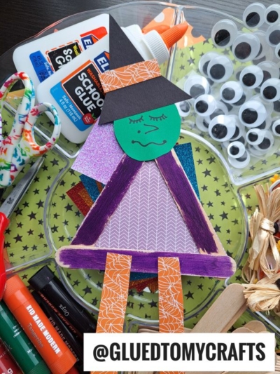 A popsicle stick graduation craft inspired by a popsicle stick Halloween witch features a green face, black cap, and purple legs, surrounded by school supplies like glue, scissors, googly eyes, markers, and colored paper. @GLUEDTOMYCRAFTS is at the bottom.