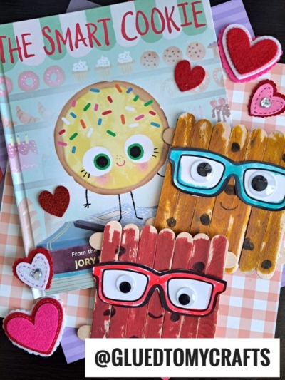 A children’s book titled The Smart Cookie with a smiling cookie on the cover is surrounded by heart decorations and two Popsicle Stick Smart Cookie crafts with large eyes and glasses. The handle @GLUEDTOMYCRAFTS is at the bottom.