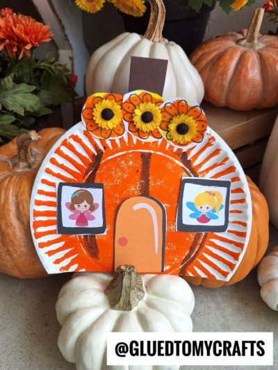 A paper plate pumpkin fairy house craft decorated with orange paint, flowers, a door, and two drawings of girls, surrounded by real pumpkins. The image has the text @GLUEDTOMYCRAFTS at the bottom.