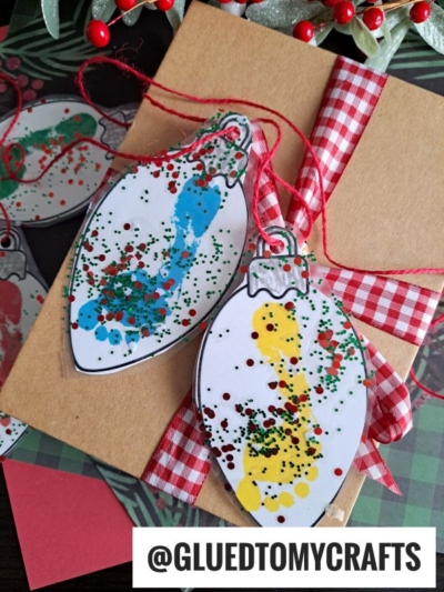Two handmade Christmas light bulb ornaments with child’s painted fingerprints and glitter are tied with red ribbon on a brown card, featuring a red gingham bow—perfect to pair with a Footprint Christmas Ornament Garland. The @GLUEDTOMYCRAFTS handle is at the bottom.