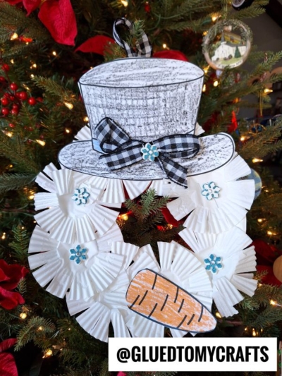 A snowman Christmas tree ornament made from white paper cupcake liners, a black paper hat with a plaid bow, and blue flower decorations, just like those on a festive snowman wreath, hangs on a beautifully decorated tree.