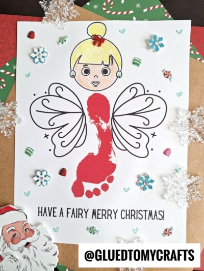 A Footprint Christmas Fairy craft with a cartoon fairy head, hand-drawn wings, and a red painted footprint body. The paper features snowflakes, hearts, and candy canes. Text reads: “HAVE A FAIRY MERRY CHRISTMAS!”.