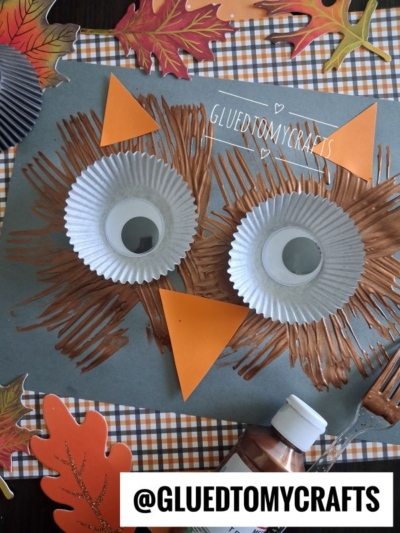 A fork painted owl craft made with brown paint, cupcake liners for eyes, googly eyes, and orange paper for ears and beak. Surrounded by fall leaves and checkered paper. A paint bottle and fork are visible in the scene.