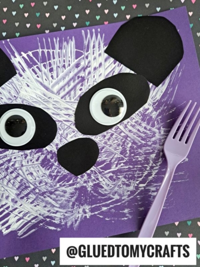 A fork painted panda face is crafted on purple paper using black paper shapes, large googly eyes, and white paint applied with a plastic fork; a lavender fork rests beside the artwork. Heart-patterned paper decorates the background.