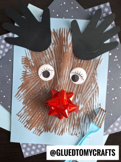 A fork painted reindeer craft made with brown strokes on blue paper, featuring large googly eyes, black paper antlers, and a shiny red gift bow nose. @GLUEDTOMYCRAFTS is written at the bottom.