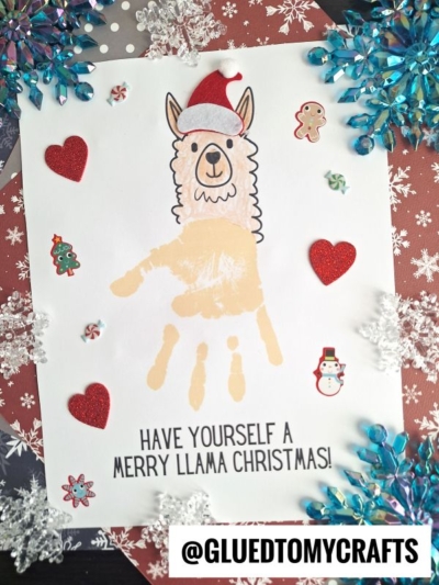A Handprint Llama Christmas craft featuring a llama drawing with a Santa hat, decorated with hearts, snowflakes, holiday stickers, and the message “Have yourself a Merry Llama Christmas!”.