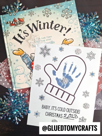 A winter-themed children’s book titled Its Winter! sits beside a mitten handprint craft featuring a blue handprint inside, snowflake decorations, and the text Baby, its cold outside! Christmas 2023 @GLUEDTOMYCRAFTS.