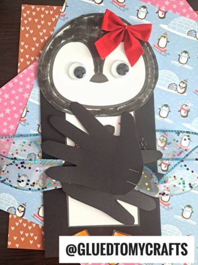 A handmade handprint penguin card featuring googly eyes, a red bow, and black paper hands as wings. The card sits on colorful penguin-themed paper with a ribbon and @GLUEDTOMYCRAFTS label at the bottom.
