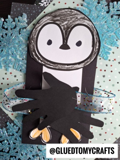 A homemade handprint penguin craft made from black and white paper, with flipper-shaped hands, yellow feet, and a polka-dot ribbon, set on glittery blue snowflakes. Text at the bottom reads @GLUEDTOMYCRAFTS.