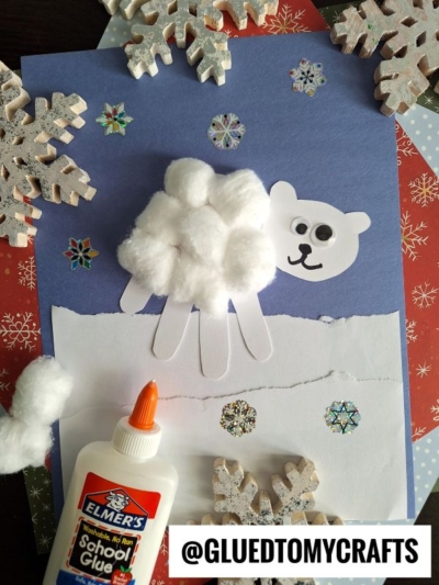 Childrens craft of a polar bear using cotton balls, cut-out paper, and googly eyes on a blue background. Snowflakes and an Elmers glue bottle are nearby—perfect for christmas keepsakes. Instagram handle @GLUEDTOMYCRAFTS is visible.