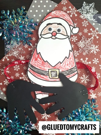 A handmade handprint Santa hug craft featuring a colored paper Santa with googly eyes, black paper hand cutouts as mittens, and a red glittery pipe cleaner for arms. Snowflake decorations and patterned paper complete the festive background.