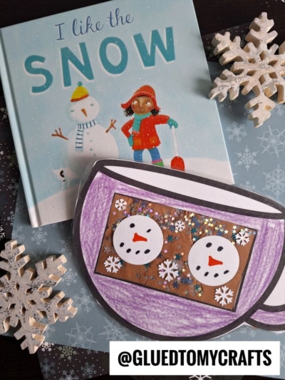 A children’s book titled I Like the Snow sits next to a Hot Cocoa Mug Craft featuring marshmallow snowmen in a cup, glitter, and paper snowflakes. The image includes the Instagram handle @GLUEDTOMYCRAFTS.