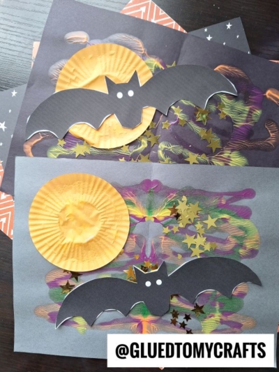 Two Halloween crafts featuring black paper bats with white eyes, yellow cupcake liner moons, and gold star confetti on dark, colorful painted backgrounds create a whimsical scene. This paint splat bat design is perfect for adding a spooky touch to your holiday decor. @GLUEDTOMYCRAFTS is written at the bottom.