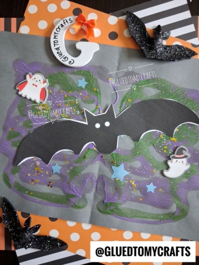 A Halloween paint splat bat craft with colorful paint, glitter, ghost and witch stickers on paper. Black and white striped and polka dot borders frame the scene, accented by @gluedtomycrafts tags. Two paint splat bat cutouts and small star doodles decorate this festive setup.