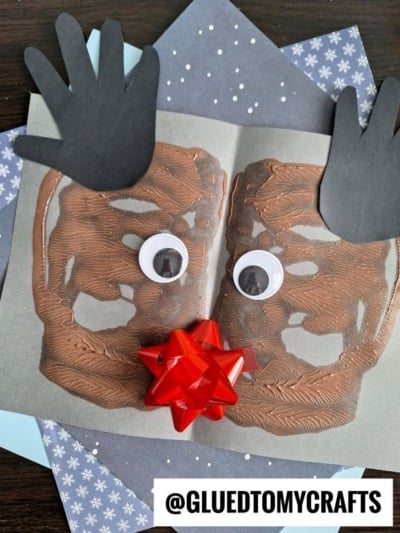 A handmade paint splat reindeer craft features a brown painted face, black paper handprint antlers, googly eyes, and a red bow nose on gray paper with a snowy background.