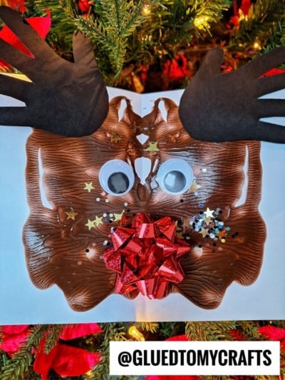 A paint splat reindeer made from brown paint, black paper antlers, googly eyes, a red bow for the nose, star-shaped confetti, and glitter sits in front of a decorated Christmas tree. @GLUEDTOMYCRAFTS is written at the bottom.