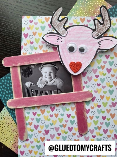 A cute reindeer picture frame craft featuring a pink reindeer with googly eyes and a red heart nose, above a pink popsicle stick frame holding a black-and-white photo of a smiling child, set on a colorful heart-patterned background.