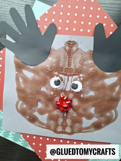 A paint splat reindeer face craft featuring brown paint blots, black paper handprints as antlers, googly eyes, and a red bow for the nose, all arranged on layered festive paper. Text: @GLUEDTOMYCRAFTS.