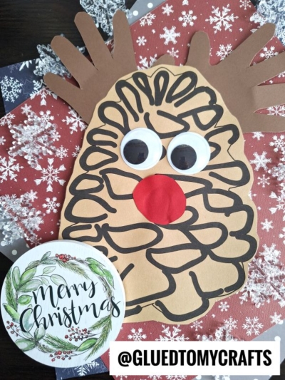A charming pinecone reindeer craft with googly eyes and a red nose is placed on snowflake-patterned paper. A Merry Christmas wreath card and the text @GLUEDTOMYCRAFTS are also visible.