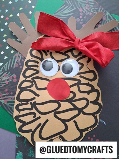 A handmade reindeer craft, inspired by a classic pinecone reindeer, features brown paper with black marker lines, large googly eyes, a red nose, antlers, and a red ribbon bow, all displayed on festive holiday paper. @GLUEDTOMYCRAFTS is written at the bottom.