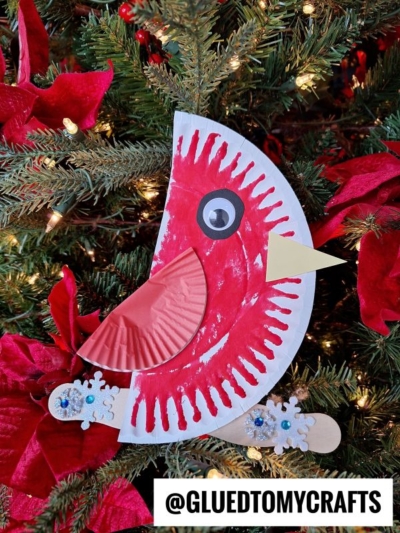 A charming paper plate cardinal craft, featuring a painted paper plate, a cupcake liner wing, and a paper beak, is displayed on a Christmas tree adorned with poinsettias and twinkling lights. @GLUEDTOMYCRAFTS text appears at the bottom.