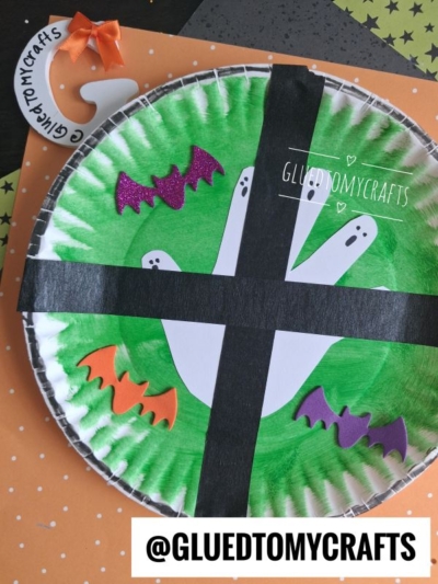 A paper plate colored green with a white handprint ghost, decorated with purple and orange bat cutouts and black tape forming a ghost window craft cross, on a polka dot background. @GLUEDTOMYCRAFTS tag is visible.
