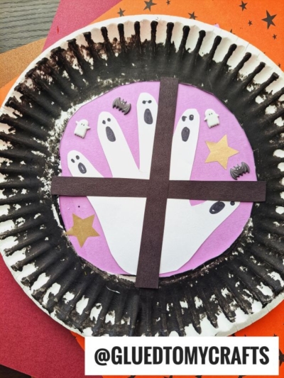 This ghost window craft features a black-painted paper plate with a purple center, white handprint ghosts, black paper strips as a window, gold stars, and cute ghost and bat stickers. @GLUEDTOMYCRAFTS is written at the bottom.