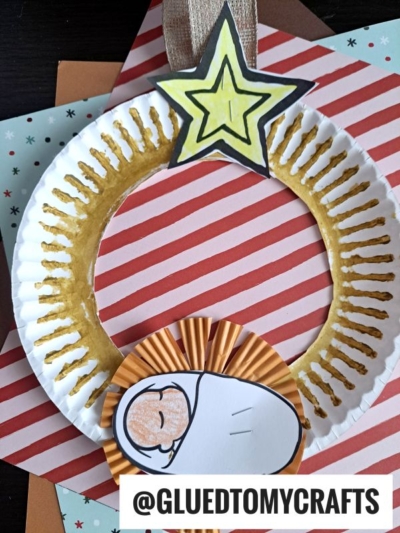A Paper Plate Nativity Wreath decorated with gold paint and a yellow star on top, featuring a baby Jesus made from paper and a cupcake liner. Red striped and star patterned papers form the background. @GLUEDTOMYCRAFTS is written at the bottom.