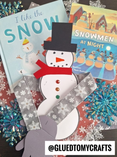 A snowman craft made of paper is displayed with two children’s books about snow and snowmen, set on a table decorated with snowflake ornaments. The setup radiates a cozy snowman hug. Image includes the watermark @GLUEDTOMYCRAFTS.
