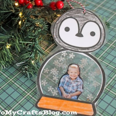A handmade Penguin Snow Globe Ornament displays a child's photo inside the globe with snowflake glitter, set on a green plaid surface beside pine branches.