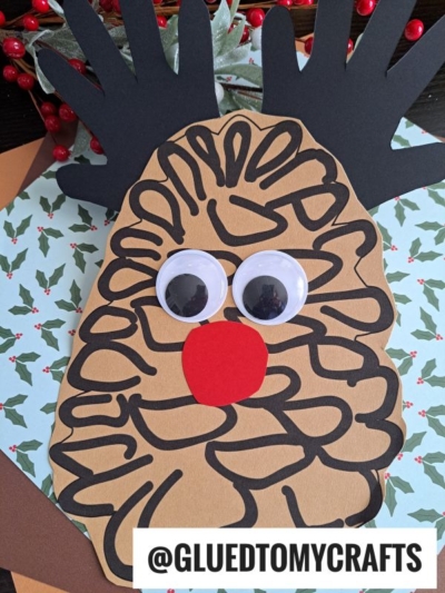 A handmade pinecone reindeer craft featuring brown paper shaped like a pinecone with black marker lines, big googly eyes, a red nose, and black paper antlers. Holly-patterned paper and red berries add festive charm in the background.