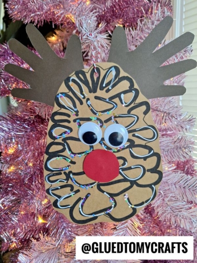 A handmade pinecone reindeer ornament with googly eyes, a red nose, brown paper handprint antlers, and glittery black swirls hangs on a pink Christmas tree. Text at the bottom reads @GLUEDTOMYCRAFTS.