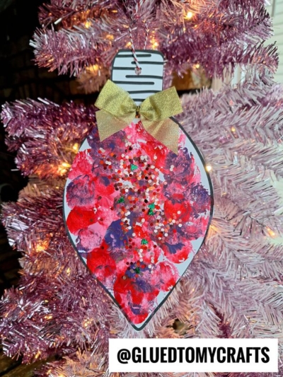 Handmade pom pom stamped ornament shaped like a bauble, painted red and purple, decorated with multicolored glitter and a gold bow, hanging on a pink Christmas tree. The tag @GLUEDTOMYCRAFTS appears at the bottom.