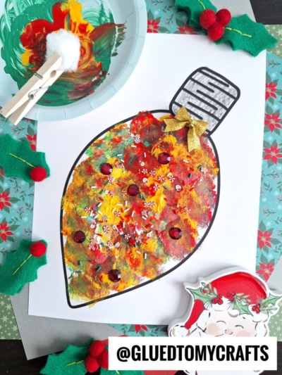 Colorful painted Christmas light bulb craft with red, yellow, and white paint, gold glitter, and decorated with small embellishments, on festive paper. A pom pom stamped ornament adds extra charm to the scene, while a clothespin with cotton ball and felt holly leaves are nearby.