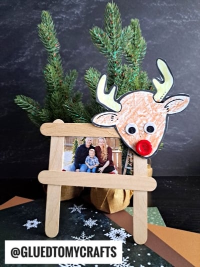 A reindeer picture frame made from popsicle sticks and featuring a family photo stands in front of a small pine tree, decorated with a handmade reindeer face. Craft materials and the handle @GLUEDTOMYCRAFTS are visible.