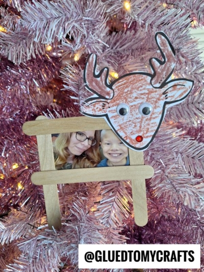 Handmade reindeer picture frame ornament hangs on a pink Christmas tree. It features a child's drawing of a reindeer with googly eyes, plus a photo of a woman and child framed with craft sticks. @GLUETOMYCRAFTS is at the bottom.