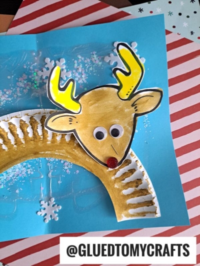 A handmade reindeer snow craft idea for kids on blue paper with white snowflakes, featuring a painted paper plate, yellow antlers, googly eyes, and a red glitter nose. The image includes the handle @GLUEDTOMYCRAFTS at the bottom.