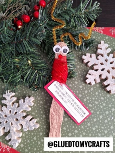 A handmade Christmas craft features a wooden spoon decorated as a reindeer keepsake with googly eyes, a red nose, red scarf wrap, gold pipe cleaner antlers, and is surrounded by holiday greenery and two wooden snowflakes, labeled @GLUEDTOMYCRAFTS.
