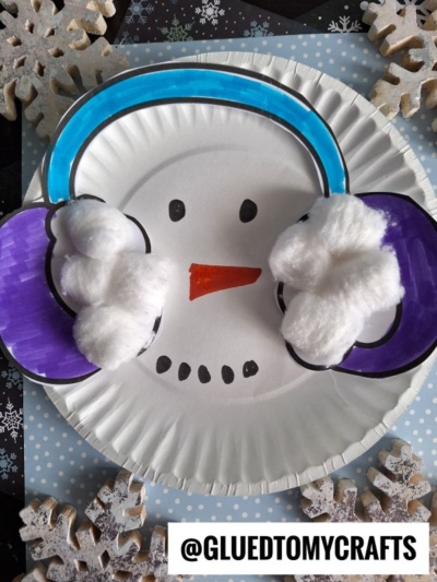 This adorable paper plate snowman craft features blue and purple earmuffs made from markers and cotton balls, with a drawn orange carrot nose and black eyes and mouth, all set against a blue background decorated with snowflakes. Perfect for winter fun, this paper plate snowman brings seasonal cheer to any crafting day!.