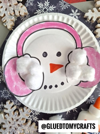 A paper plate snowman features pink cotton ball earmuffs, a drawn orange carrot nose, and small black eyes on a snowflake-patterned background. A glue bottle and white snowflakes complete this adorable paper plate snowman craft.