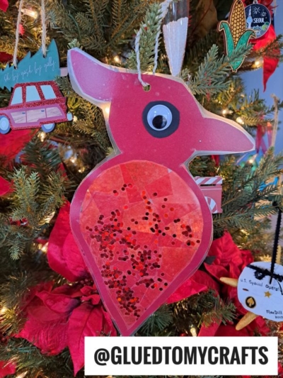 A handmade red bird ornament, reminiscent of a cardinal suncatcher, with a googly eye and glitter, hangs on a Christmas tree decorated with flowers and various ornaments. @GLUEDTOMYCRAFTS is written at the bottom of the image.