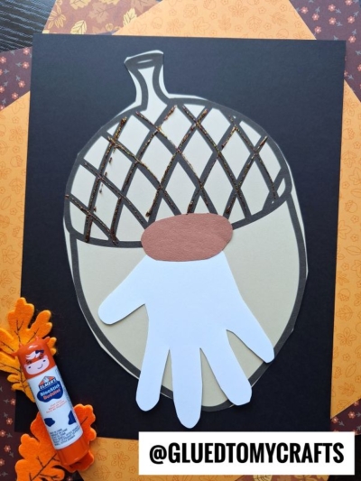 A paper craft acorn gnome features a charming white handprint as its base, placed on a striking black background. A glue stick, felt leaves, and patterned paper are arranged nearby. The text @GLUEDTOMYCRAFTS appears at the bottom.