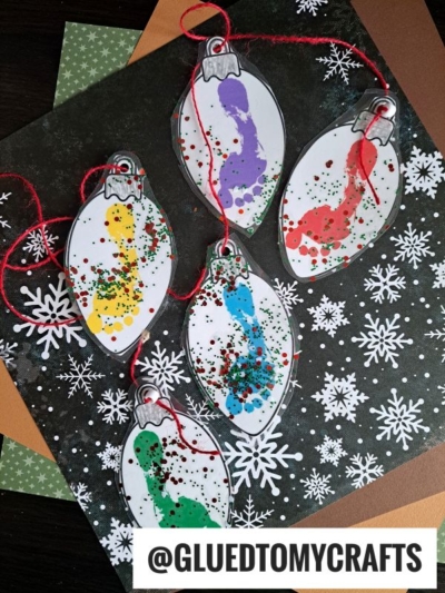 Five paper Christmas light-shaped ornaments, perfect for a Footprint Christmas Ornament Garland, feature colorful paint footprints and glitter. Displayed on snowflake-patterned backgrounds, each has red hanging strings. @GLUEDTOMYCRAFTS is written at the bottom.