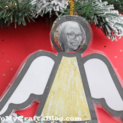A handmade angel photo ornament features a black-and-white picture of a smiling woman as the face, colored yellow and white, hanging before pine branches.