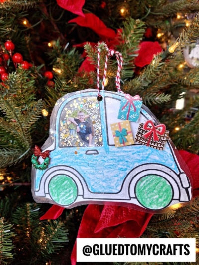 A handmade blue christmas car ornament colored with crayons hangs on a Christmas tree. The car has gifts on top, a photo in the window, and festive bow and holly decorations. @GLUEDTOMYCRAFTS is written at the bottom.