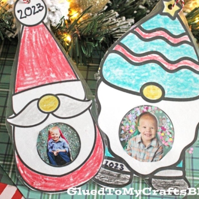 Two Christmas Gnome Photo Ornaments labeled “2023” feature cut-out circles for children’s photos. Resting on a plaid background with pine branches