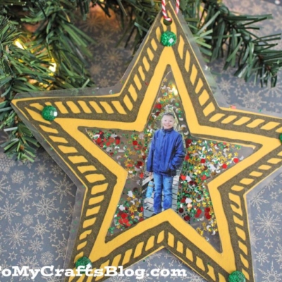 A christmas star ornament with a photo of a child in the center hangs on the tree. The yellow ornament, decorated with black details and green dots, stands out among the festive lights and branches in the background.
