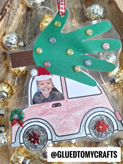 A handmade christmas car ornament showcases a smiling child in a pink paper car with festive wheels, carrying a green handprint tree. Surrounded by string lights and gold ornaments. Text: @GLUEDTOMYCRAFTS.