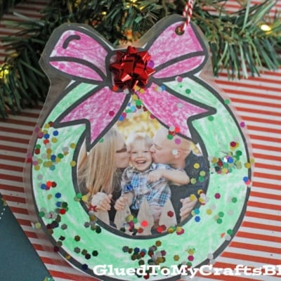 A handmade wreath photo ornament shaped like a Christmas wreath with a pink bow, glitter accents, and a photo of a child being kissed by two adults.