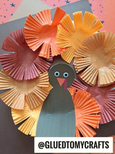 A cupcake liner turkey craft made with colorful orange, red, and yellow cupcake liners for feathers, a brown paper body with googly eyes and a beak, displayed on layered paper. @GLUEDTOMYCRAFTS is written in the corner.